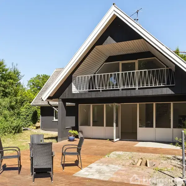 Sort træhus med A-formet tag har en stor terrasse og havemøbler foran. Huset har en balkon på første sal. Det er omgivet af grønne træer og en blå himmel.