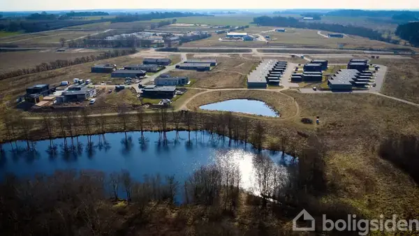 En stor sø reflekterer lys, omgivet af træer. Bagved ligger en række bygninger, spredt i et åbent område. I baggrunden ses marker og spredte skove.