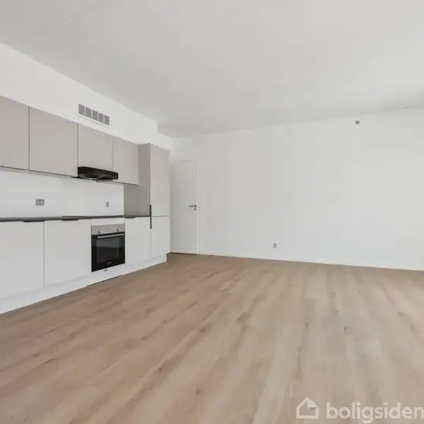 Et køkken med indbyggede skabe og apparater, herunder en ovn og emhætte, står i et lyst, tomt rum med lyse trægulve og hvide vægge, skaber en ren og moderne atmosfære.