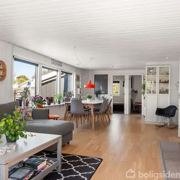 Grå sofa står ved et sofabord med planter ovenpå. I baggrunden ses et spisebord med stole, belyst af en rød lampe. Rummet har hvide vægge og trægulv.