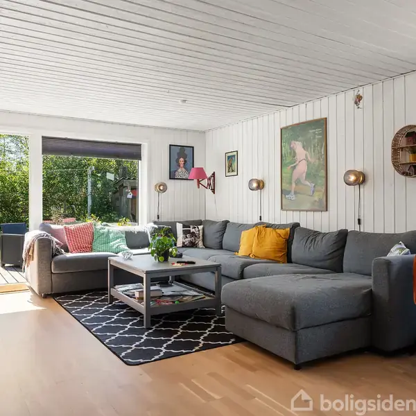 En stor, grå sofa står i en stue oplyst af naturligt lys fra en åben dør til haven. Hyggelige puder og tæpper, en sort-hvidt tæppe, og moderne kunst pynter rummet.