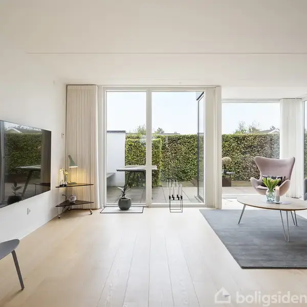 En moderne stue har en sofa og lænestol på et tæppe. Et stort vindue åbner mod en terrasse med grønne hække. Enkel indretning med mørke møbler og lys trægulv.