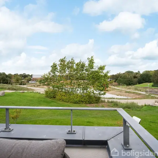 Terrasse med gelænder; ser ud over en grøn have. En person går med en hund i baggrunden. Skyet himmel og spredte træer omkranser området.