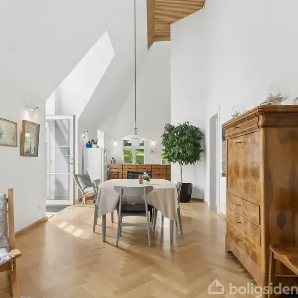 En spisestue har et bord med stole under en lampeskærm. Rummet er lyst og rummer møbler som en skænk og stol. Kunst og en plante præger hvide vægge.