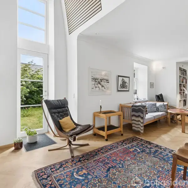 En lys stue med høje glasdøre viser et moderne rum. En lænestol står på et orientalsk tæppe ved siden af en sofa med ryatæppe. Grøn udsigt udenfor.