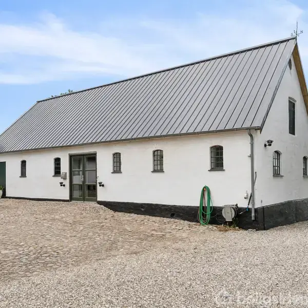 En hvid bygning med grått tag står på en brolagt gårdsplads. En haveslange hænger på væggen. Der er åbent landskab og træer i baggrunden under en blå himmel.