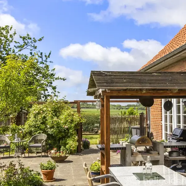 Terrasseområde med havemøbler og grill under en pergola. Omgivet af blomster, træer og en grøn have med udsigt til det åbne landskab og en blå himmel med skyer.