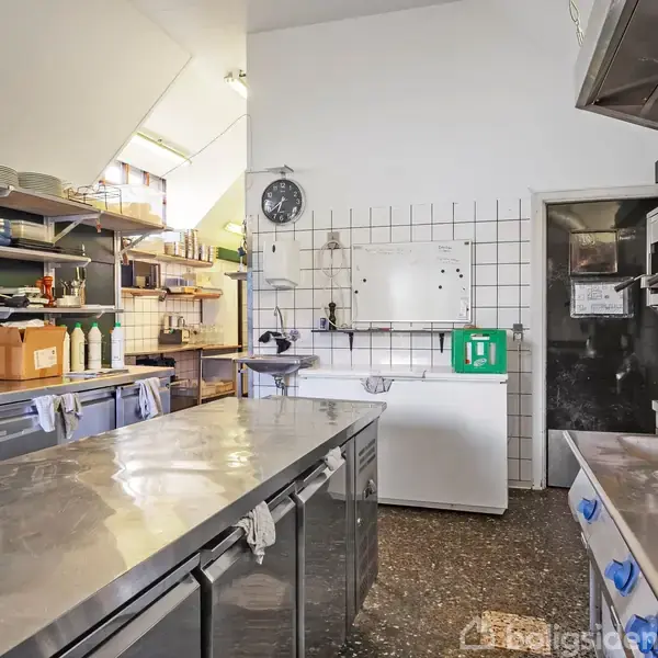 Stålkøkkenbord med redskaber står i et industrielt køkken med hvide fliser, hylder med tallerkener og en stor ovn. Rummet er oplyst af to vinduer øverst til venstre.