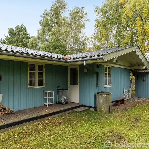 Et blåt træhus med hvidt tag står stille i en grøn have omkranset af træer. Foran huset ligger en lille veranda med brændestabel og hvid bænk.