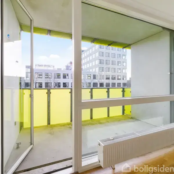 En stue med et åbent vinduesparti, der fører ud til en altan med gule skærme. Udsigt til flere moderne bygninger udenfor. Parketgulv og radiator indendørs.