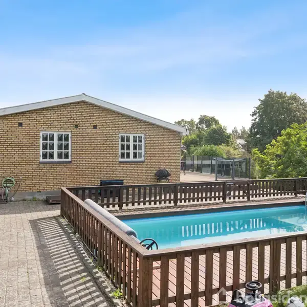 En rektangulær swimmingpool omgivet af et træhegn står stille på en flisebelagt terrasse foran et gult murstenshus med hvide vinduer. Træer ses i baggrunden.