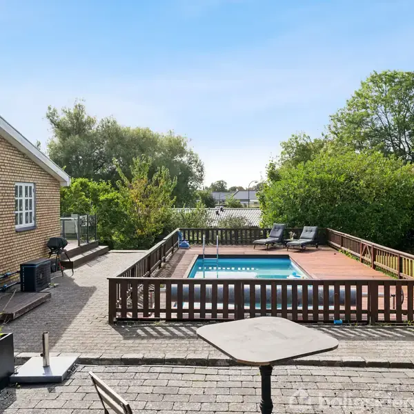 En swimmingpool er omgivet af træhegn og liggestole i en have. Brolagt terrasse med borde og stole i forgrunden, tæt på et gult hus. Grønt løv omkranser området.