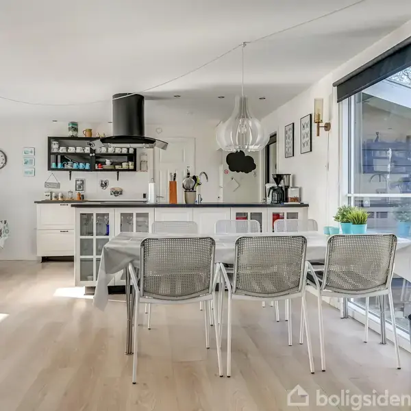 Et moderne køkken-alrum med spisebord og fire stole, en brændeovn i hjørnet, og et hvidt køkken i baggrunden. Store vinduer giver naturligt lysindfald.