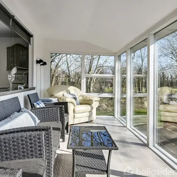 Stole i polyrattan med blå og hvide puder står i en glasindelukket veranda med udsigt til en skov. Et lyst sofahjørne kompletterer det hyggelige rum.