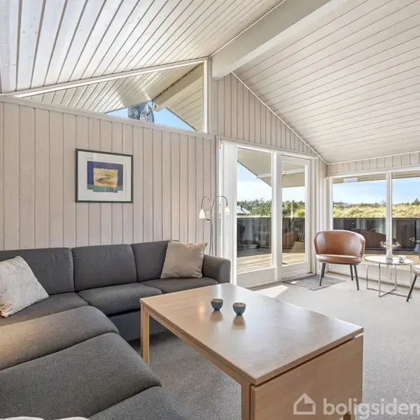 Sofaen står foran en trævæg med et billede. Stuen er lys med store vinduer mod en terrasse. To lænestole og et lille bord står ved vinduerne.