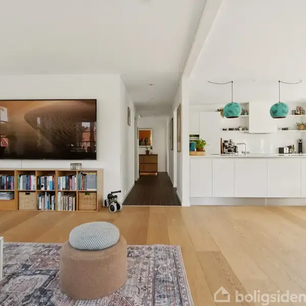 Et moderne stue-interiør med trægulv, stor fladskærms-TV på væggen, en reol fyldt med bøger, køkken med hvidt inventar, og to turkise lamper hængende over køkkenbordet.