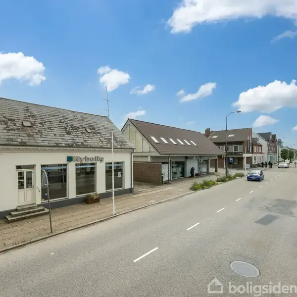 En vej ved siden af en række huse, herunder en bygning med skiltet "Nybolig". Parkerede og kørende biler ses i begge retninger under en klar blå himmel med få skyer.