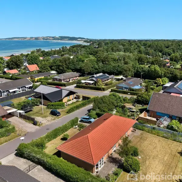 Boligområde med en række huse med røde og grå tage, omgivet af grønne haver og træer. Kystvendt landskab med udsigt over havet i baggrunden på en solrig dag.