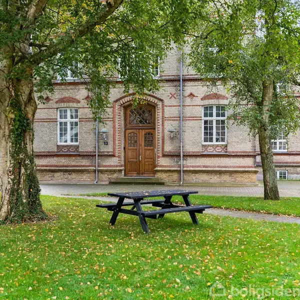 En sort bænk står stille på en græsplæne omgivet af træer. I baggrunden ses en ældre murstensbygning med en stor trådør og hvide vinduer.