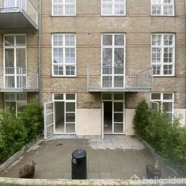 Bygning med tre etager, hver har store vinduer og balkoner. Dobbelt terrassedør er åbnet i stueetagen. Omkranset af buske, med en skraldespand foran på flisebelagt terrasse.