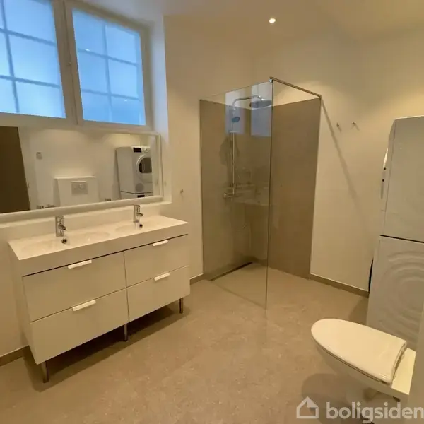 Toilet i moderne badeværelse med dobbeltvask og spejl; brusebad med glasvæg ved siden af. Vaskemaskine og tørretumbler er synlige. Lyse fliser på væg og gulv.