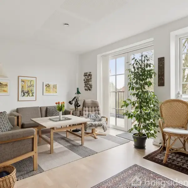 Stue med sofaarrangement og sofabord, omgivet af indrammet kunst på væggen. Store vinduer giver udsigt til træer, mens planter og kurvestole skaber en hyggelig atmosfære.