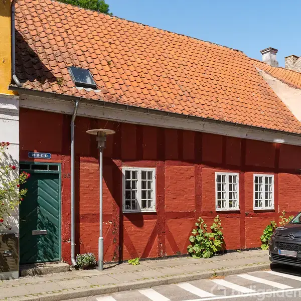 Et rødt murstenshus med orange tegltag og grøn dør står ved en gade. Huset har hvide vinduer, og en bil er parkeret foran. Adresse-skiltet viser "26C".