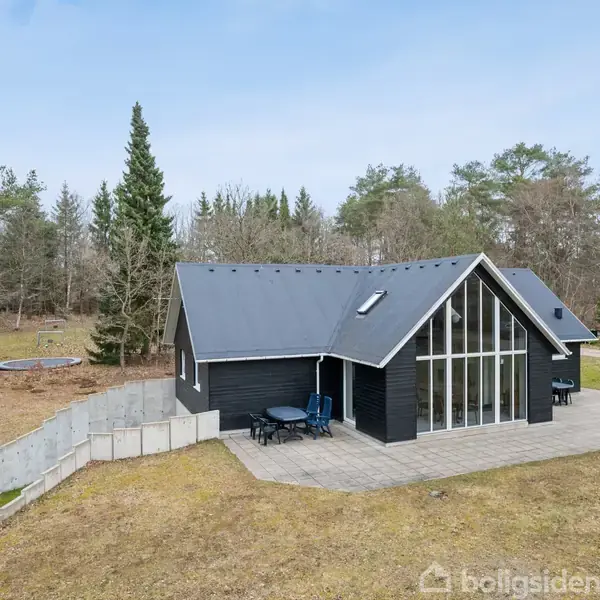 Sort træhus med store glaspartier står stille på en flisebelagt terrasse omgivet af en græsplæne med træer i baggrunden.