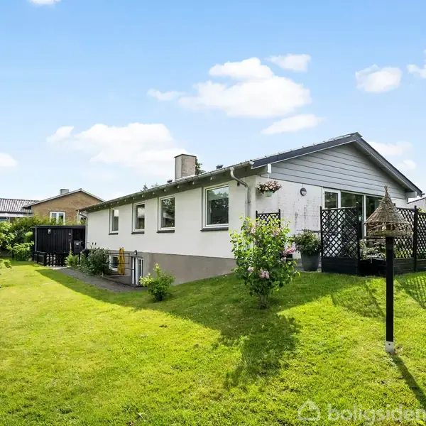 Et enfamiliehus står på en velplejet græsplæne. Huset er hvidt med gråt tag, omgivet af planter og haveelementer. Himmel er blå med få skyer.