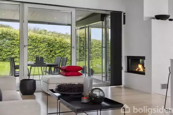 Stue med moderne indretning og brændende pejs. Gennem store glasdøre ses en terrasse med møbler og en grøn hæk udenfor. Røde puder ligger på en hvid bænk.