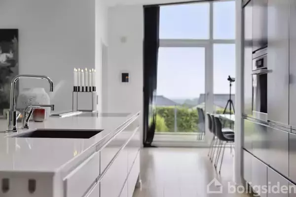 Køkken med hvidt moderne design, hvor en vandhane står på en blank køkkenø. Store vinduer slipper lys ind, udsyn til grønne træer. Ingen synlig tekst bemærket.