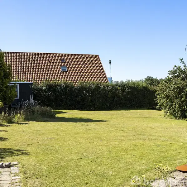 Græsplæne udstrakt over en have, indrammet af buske og et hus med rødt tegltag i baggrunden. En træbænk står til højre. Himmelstrækning er klar og blå.