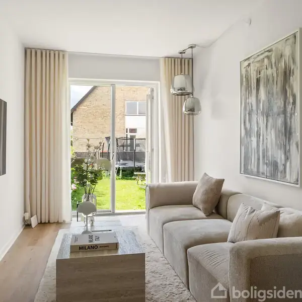 Stue med lys sofa står foran en stor skydedør, der åbner til en have. Et moderne maleri hænger på væggen, og bøger ligger på et sofabord.