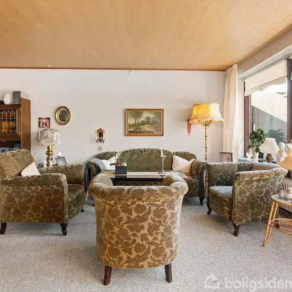 En stue med retro møbler består af blomstrede lænestole og en sofa, arrangeret rundt om et sofabord. En bogreol og en standerlampe ved siden af et vindue skaber en hyggelig atmosfære.