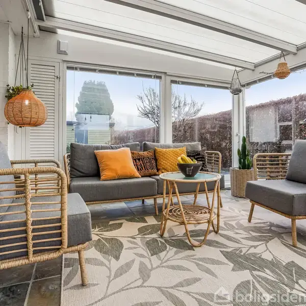 Lyst møbleret haveværelse med kurvemøbler og grå hynder. To orange puder på sofaen. Et lille rundt bord bærer en skål. Store vinduer viser en hæk udenfor.