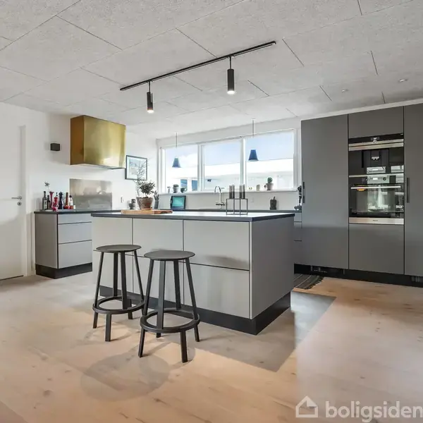 Et moderne køkken med en grå køkkenø med to sorte barstole. Skabe og indbyggede hvidevarer i gråt. Rummet er lyst med trægulv og store vinduer i baggrunden.