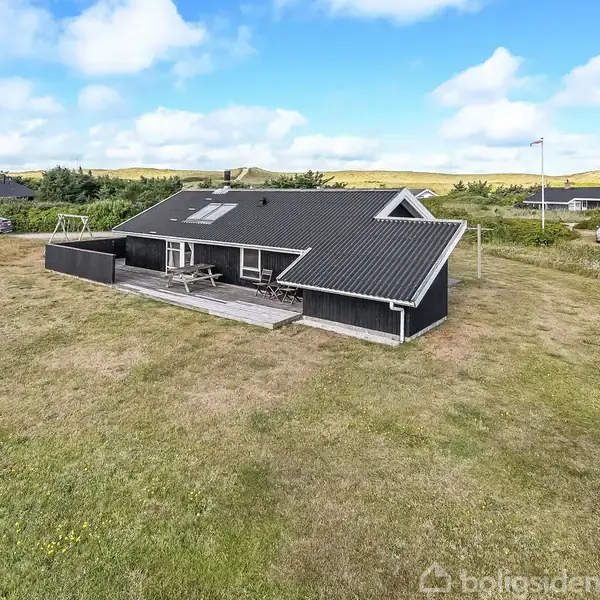 Sort træhus står stille på en åben græsplæne med blå himmel i baggrunden. En terrasse med bord og bænke er foran huset, omgivet af andre huse og grønne buske.
