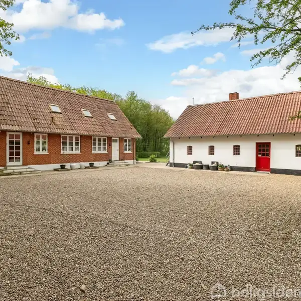 To huse står på en grusbelagt gårdsplads; det ene er rødt mursten med skråtag, og det andet er hvidt med rødt tag og dør. Omgivet af træer.
