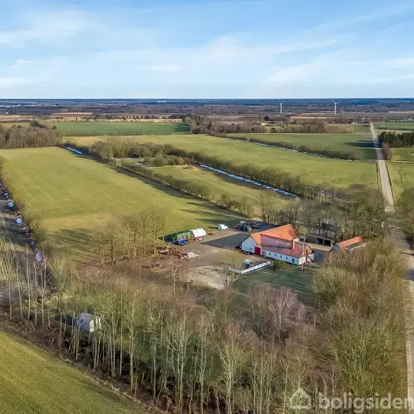 En gård med rødt tag ligger centralt, omgivet af marker og træer. Lange grusveje strækker sig gennem det åbne landskab under en blå himmel.
