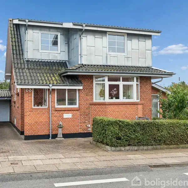 Rødt murstenshus, to etager, med kvisttag; garage ved siden. Forhaven har grønne buske. Beliggende i et boligområde med træer og blå himmel i baggrunden.