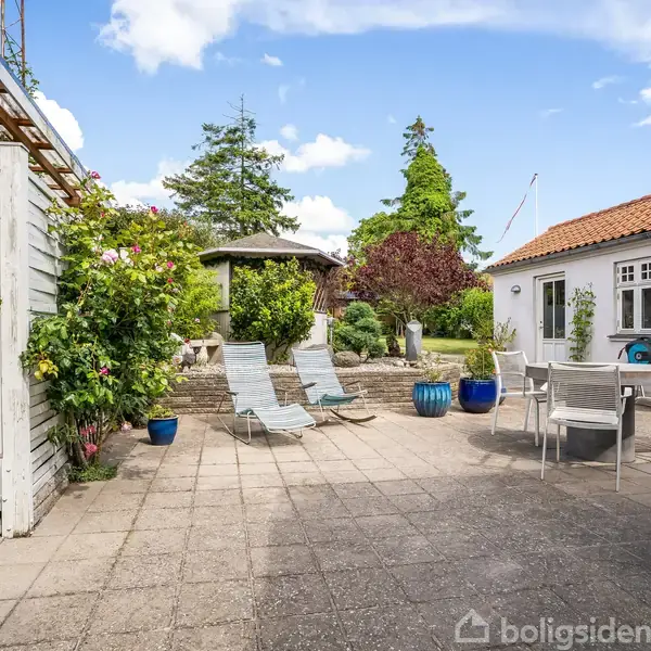 En terrasse med to liggestole og et bord med stole, omgivet af frodig beplantning og et hvidt hus med rødt tag i baggrunden under en blå himmel.