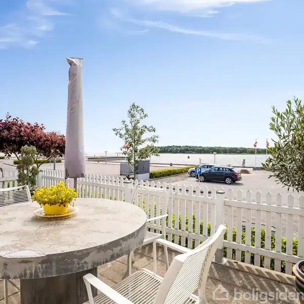 Rundt havebord med gule blomster står på terrasse omgivet af hvide havestole. I baggrunden er der en hvid stakit, en vej, parkerede biler og en udsigt over vandet.