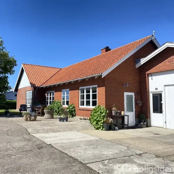 En rødstensbygning med rødt tegltag står stille omgivet af en gårdsplads. Planter dekorerer facaden ved siden af indgangen. Sollys belyser området under en klar blå himmel.