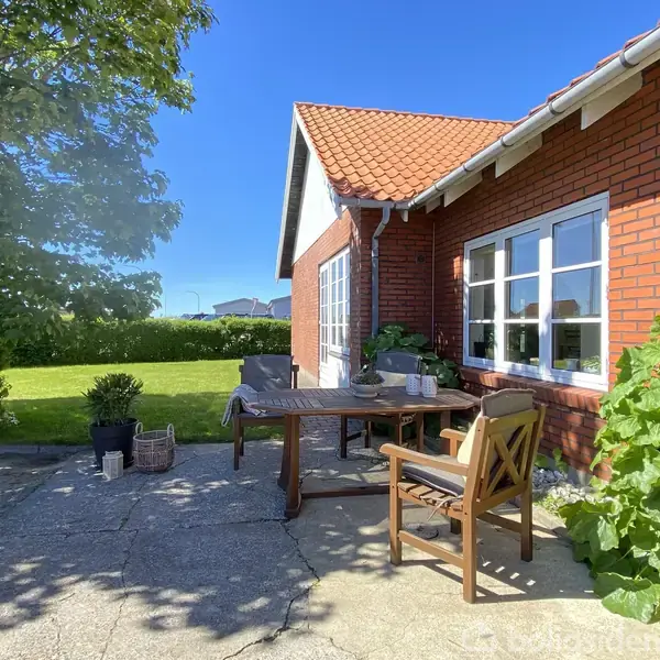 Rød murstensbygning med hvide vinduer har en terrasse med træmøbler og planter. I baggrunden ses en grøn have og en blå himmel.