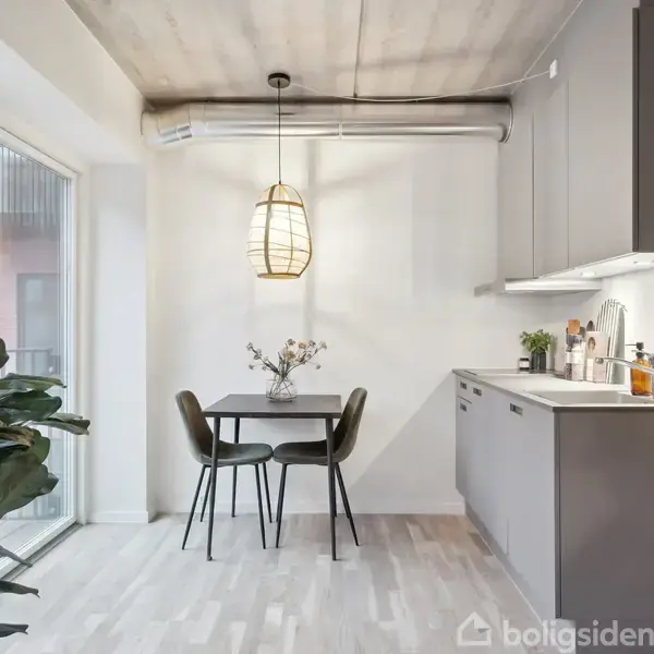 Et lille spisebord med to stole står ved siden af et moderne køkken i et lyst, minimalistisk rum. En stor plante og store glasvinduer giver naturligt lysindfald.