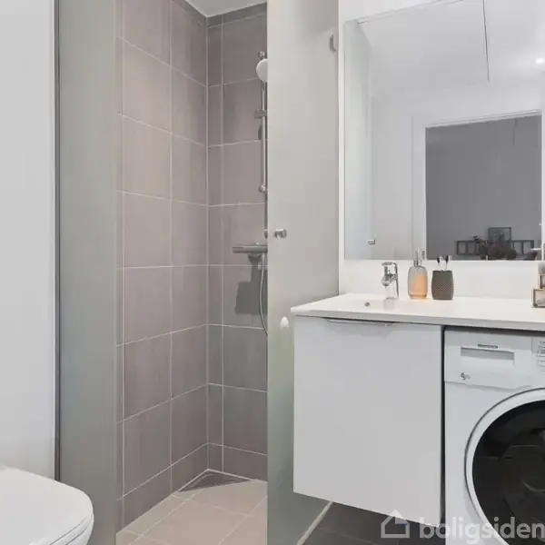 Vaskemaskine i et moderne badeværelse placeret under en hvid bordplade med spejl. Ved siden af er en bruser og et toilet. Flasker og dekorationer står på bordet.