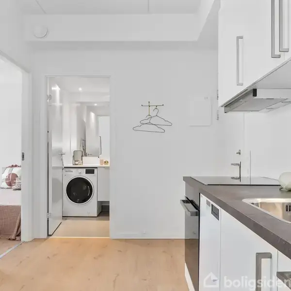 Et moderne køkken med hvidlakerede skabe og en vask på en mørk bordplade. I baggrunden er der en døråbning, der fører til et bryggers med en vaskemaskine.