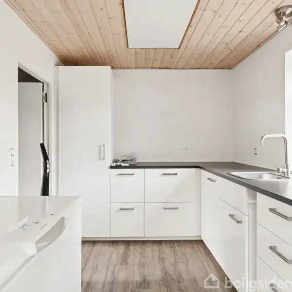 Køkken med hvide skabe og skuffer, sort bordplade og vask. Trægulv og loft. Vinduet til højre giver naturligt lys. Døråbning til venstre. Ingen synlig tekst.