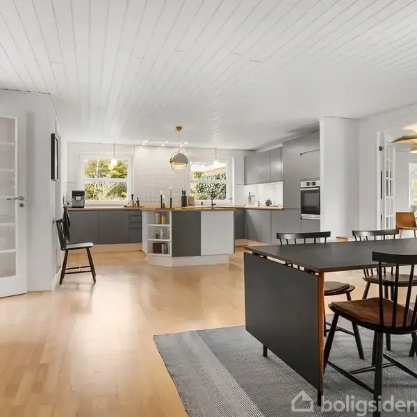 Et lyst, åbent køkkenalrum med trægulv. En spisebordssætning af mørkt træ står til højre nær en grå løber. I baggrunden ses et L-formet køkken med grå skabe og vinduer.