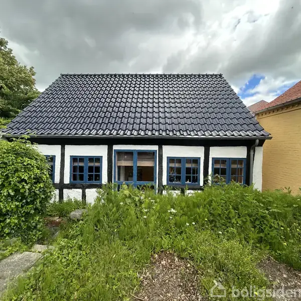En traditionel bindingsværkshus med sort tegltag og blå vinduesrammer står stille. Omgivet af en have med højt, grønt græs. Himlen er overskyet i baggrunden.